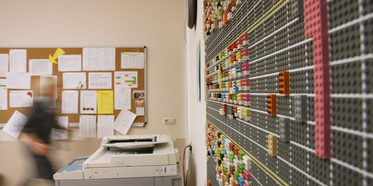 Ein überdimensional großer Stundenplan aus Legosteinen und ein Schwarzes Brett hängen an den Wänden im Lehrerzimmer der Friedensschule Schwerin. Eine Lehrerin geht zu einem Kopiergerät, das in der Ecke steht. Foto: Silke Winkler