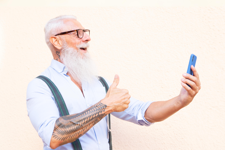 Senior, der nach rechts schaut, ein Smartphone in der linken Hand hält, den rechten Daumen nach oben zeigt und lächelt.