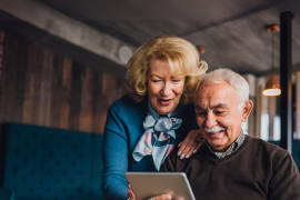 Ein Senior hält ein Tablet in der Hand und schaut lachend darauf. Hinter ihm steht eine Seniorin, die Hand auf seine Schulter gelegt und leicht nach vorne gebeugt und schaut ebenfalls lächelnd auf das Tablet. Die beiden sind in einem Raum mit braunen Holzwänden- und Decke, hinter ihnen ist eine dunkelblaue Couchgarnitur.