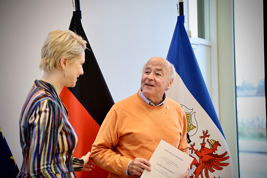 Ministerpräsidentin Manuela Schwesig mit dem Ausgezeichneten Ulrich Quast, Gründer der Initiative "Willkommen in Boizenburg". Er hält seine Urkunde in der Hand und trägt die Ehrennadel des Landes. Im Hintergrund sind die Deutschland- und die Mecklenburg-Vorpommern-Flagge zu sehen.