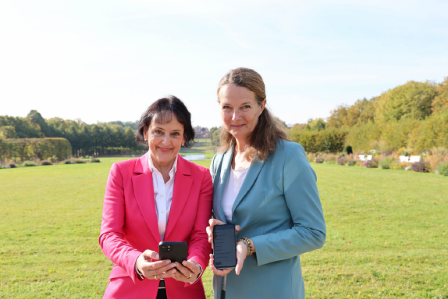 Kulturministerin Bettina Martin und die Direktorin der Staatlichen Schlösser, Gärten und Kunstsammlungen, Dr. Pirko Zinnow zeigen Handys mit der neuen App in die Kamera. Sie stehen dabei im Schlossgarten.